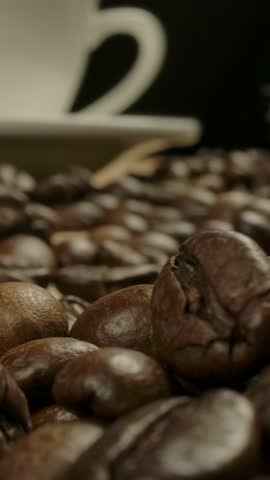 Camera moving throw roasted coffee beans to white classical coffee cup. Jute sack at the background. Wooden table, smooth dolly movement. Macro Vertical shot.