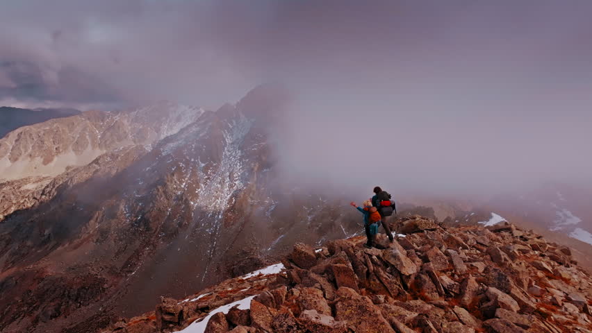 Hikers stand triumphantly on a rugged summit in the Pyrenees, surrounded by fog and majestic mountains. The view showcases the stunning landscape of northern Spain.