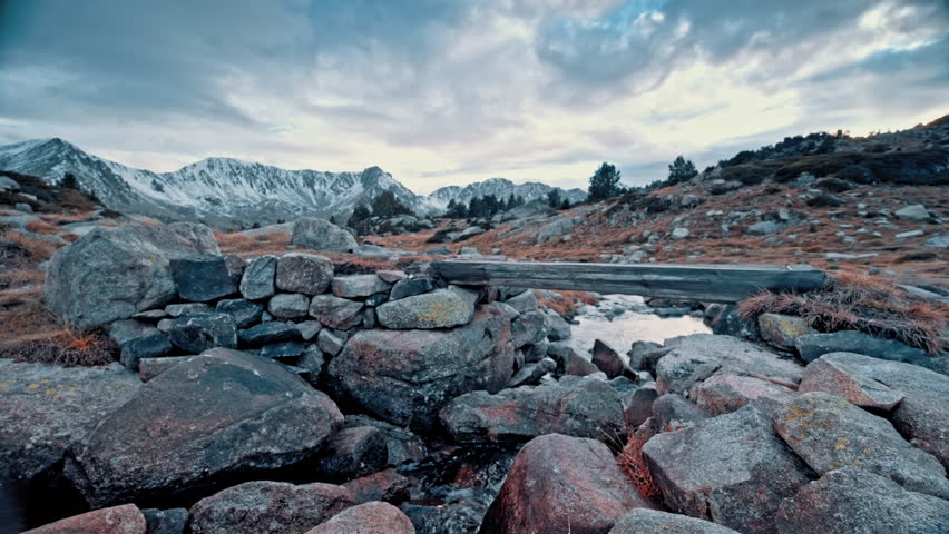 A rugged stone bridge connects two rocky banks above a gently flowing stream in the breathtaking Pyrenees mountains of Spain during the late afternoon.