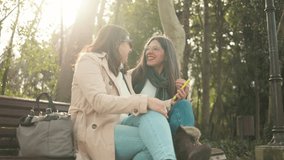Mature female friends laughing, sharing smartphone screen while sitting on park bench, enjoying warm sunlight together - Powered by Shutterstock - Get 15% off with code: PIKWIZARD15