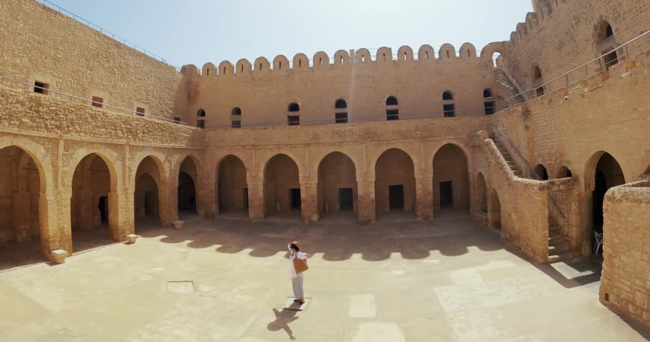 Tourist woman with wide brimmed hat rotate with arms outstretched in the courtyard of a historic Ribat fortress in Sousse, Tunisia, aerial shot