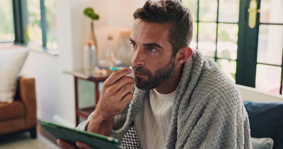 Temperature, sick and man with tablet in home for fever, influenza or cold medical symptoms. Thermometer, digital technology and male person with online telehealth consultation on sofa in living room