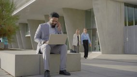 In a busy city, African American businessman sits on a bench, intently using his laptop and talking on his phone, while two women walk by, contributing to the lively urban atmosphere. Business concept - Powered by Shutterstock - Get 15% off with code: PIKWIZARD15