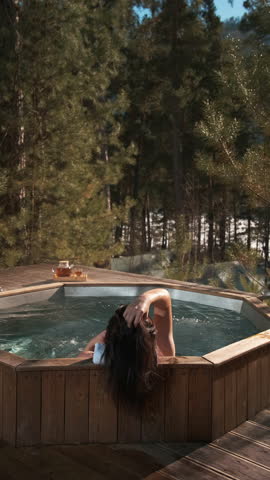 Dark haired woman relaxes in a steaming hot tub, gently moving her wet hair and enjoying the serene view of a snowy winter forest, a cup of tea placed nearby on the wooden deck