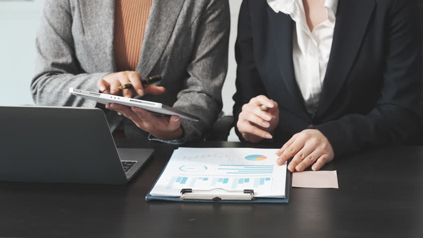 Two female business professionals in suits sit at desk, engaged in strategic discussion. They analyze financial planning, risk management, corporate growth while reviewing reports and market trends.