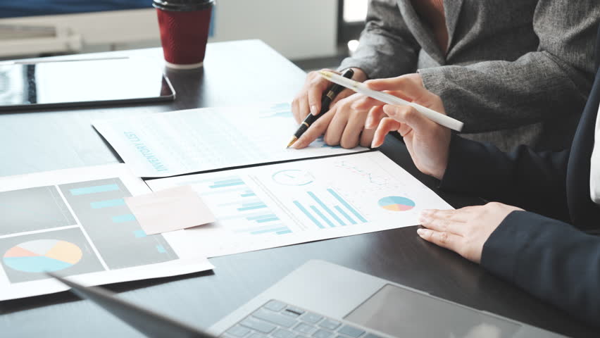 Two female business professionals in suits sit at desk, engaged in strategic discussion. They analyze financial planning, risk management, corporate growth while reviewing reports and market trends.