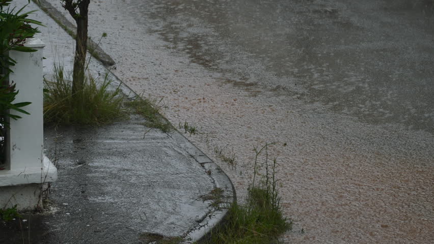 Extreme rain falls on road corner with stream of dirty water running down sidewalk. Splashes and marks on puddles from large drops of heavy rain on cloudy day with risk of flooding.