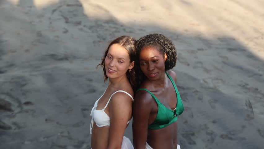Two friends pose back to back at the seaside during vacation, girls of different ethnicities