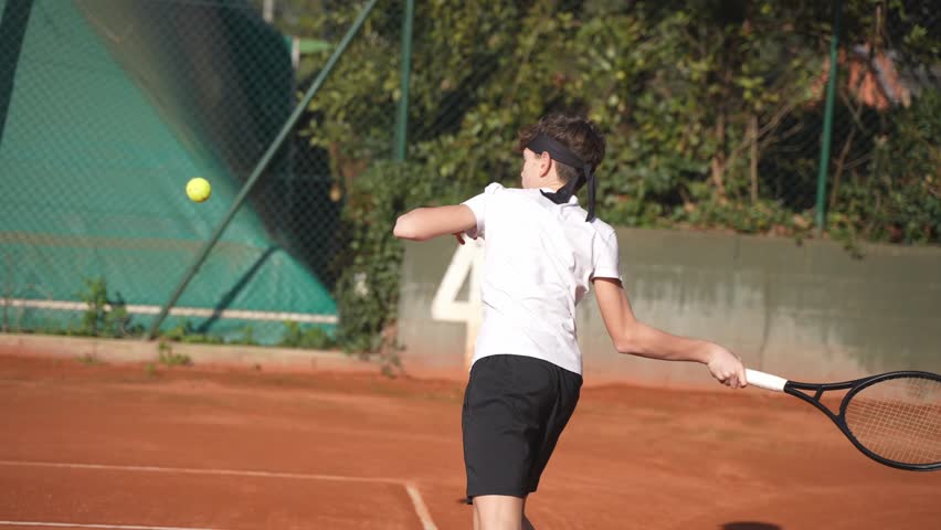 Closeup of a teenage boy with a headband playing tennis in summer on a clay tennis court.
