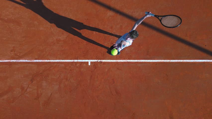 Aerial flat lay shot of a teenage male tennis player serving on a clay court.