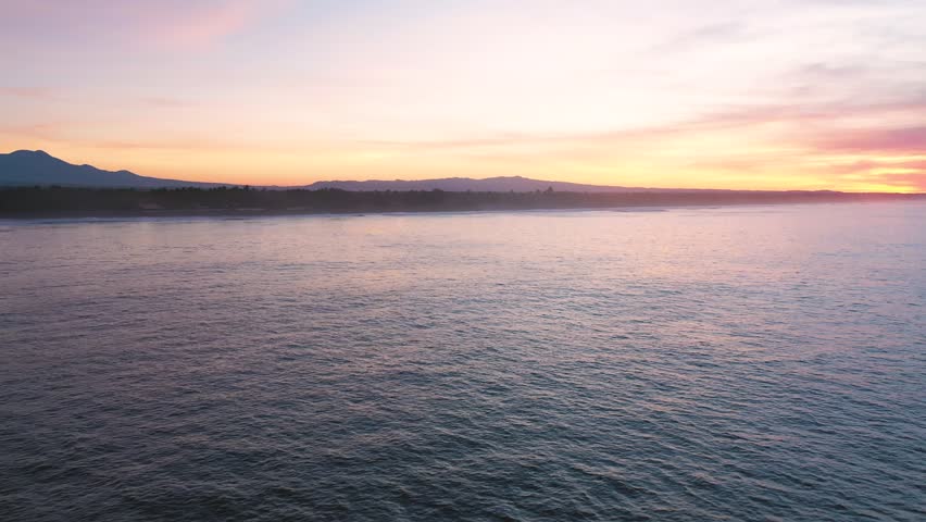 Drone flying towards the coast from the ocean. The calm water reflects the red sunset over the horizon. Aerial drone shot, 4K