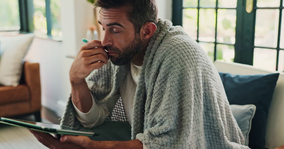 Thermometer, sick and man with tablet in home for fever, influenza or cold medical symptoms. High temperature, technology and male person with online telehealth consultation on sofa in living room.