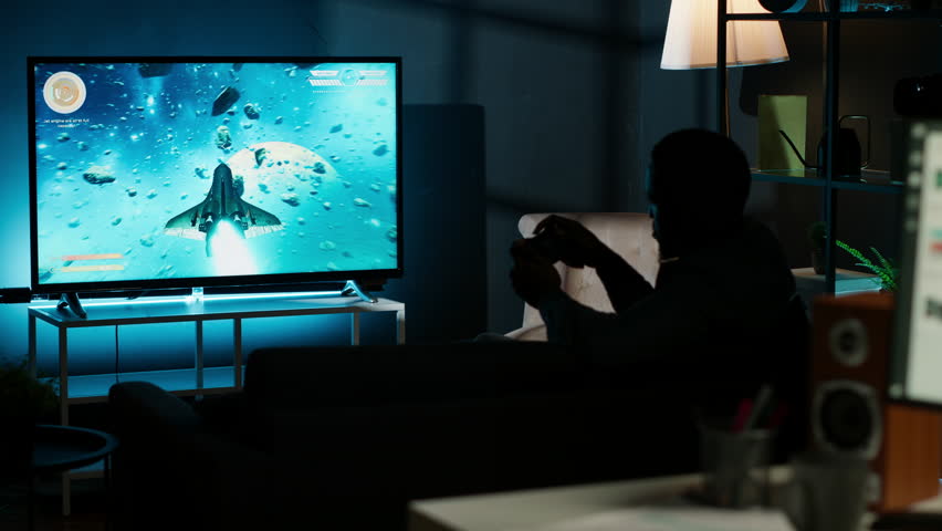 Guy spends time on television playing multiplayer web video games, setting his console into smart tv and having fun gaming at night. Player battling in shooter competition. Camera B.