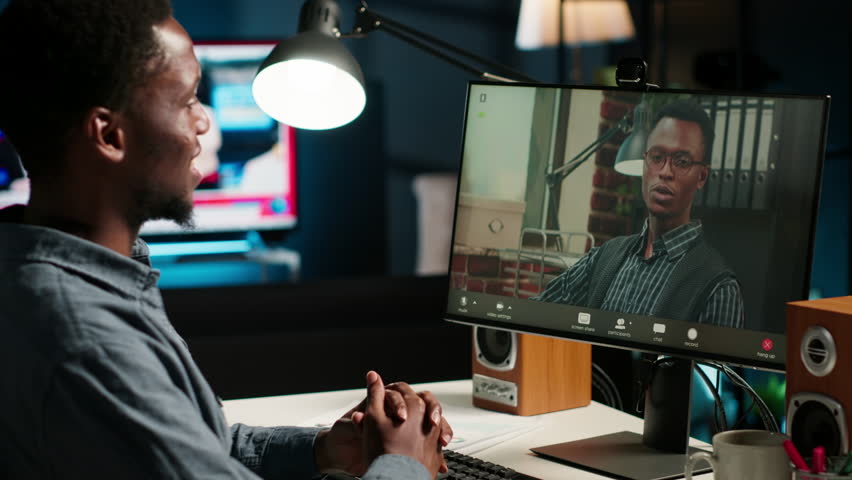 African American man enjoys video conferencing with manager at desk, discussing with his colleague online using a computer at home office setup. Young associates team meeting online. Camera B.