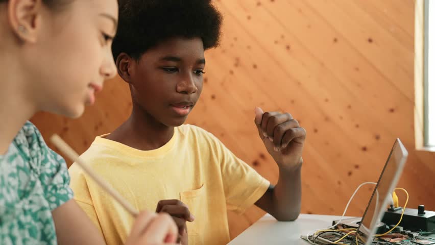 Site view of african boy and caucasian girl in casual cloth working together to coding prompt or programming system at table with laptop and electronic equipment at STEM technology class. Edification