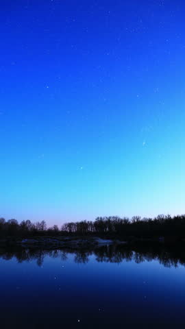 Real Colorful Night Stars Above Lake River. Natural Starry Sky Background Backdrop. Time Lapse. Vertical Shot. Glowing Stars Reflected In Lake River Water. Natural Starry Sky Background Backdrop