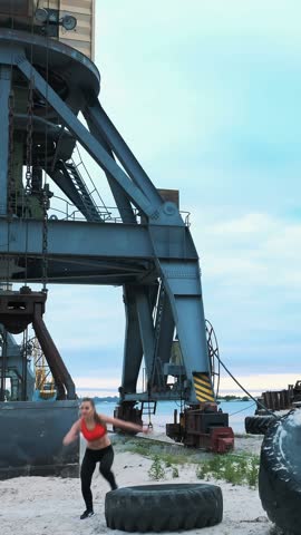 athletic woman in a red top and black leggings performs different strength exercises using a large heavy tractor wheel, trains her muscles. behind is cargo crane, cargo port, dawn