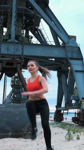 athletic woman in a red top and black leggings performs different strength exercises using a large heavy tractor wheel, trains her muscles. behind is cargo crane, cargo port, dawn
