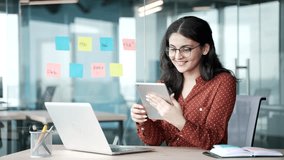 Smiling young businesswoman is using a digital tablet while sitting at a desk at a workplace in a modern business office. Happy female employee reading a message, chatting online or browsing web - Powered by Shutterstock - Get 15% off with code: PIKWIZARD15