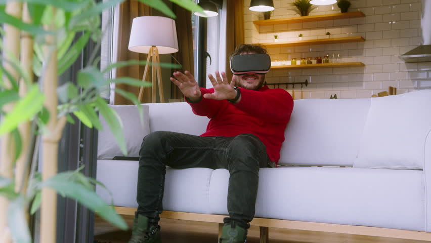 Young man wearing a VR headset, fully immersed in a virtual reality experience, gesturing with his hands while sitting on a cozy white couch in a modern living room. - Powered by Shutterstock - Get 15% off with code: PIKWIZARD15
