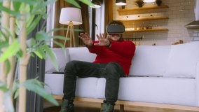 Young man wearing a VR headset, fully immersed in a virtual reality experience, gesturing with his hands while sitting on a cozy white couch in a modern living room. - Powered by Shutterstock - Get 15% off with code: PIKWIZARD15