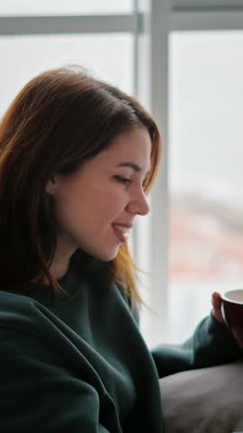 Happy brunette girl holding a cup of coffee in her hands and relaxing on the sofa at home