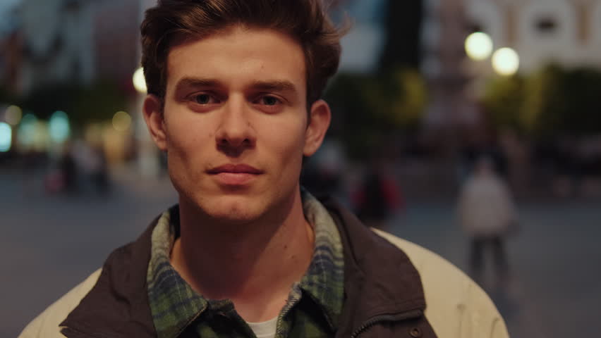 Young man stands in city square at night, turns to camera, smiles. Urban life, travel, lifestyle, portrait, male, handsome, cool, trendy, stylish