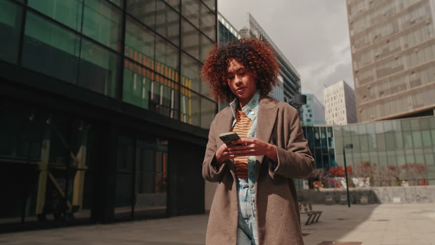 A stylish businesswoman smiles as she walks in the city while looking at her smartphone. She's happy and confident.