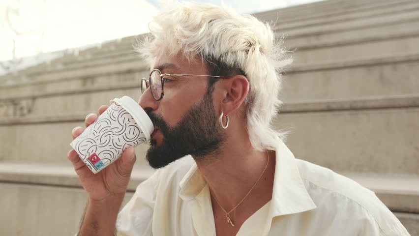Stylish man with blonde hair and beard enjoys coffee outdoors on steps. He wears glasses, a white shirt, and gold jewelry while drinking from a disposable cup.