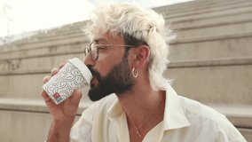Stylish man with blonde hair and beard enjoys coffee outdoors on steps. He wears glasses, a white shirt, and gold jewelry while drinking from a disposable cup. - Powered by Shutterstock - Get 15% off with code: PIKWIZARD15