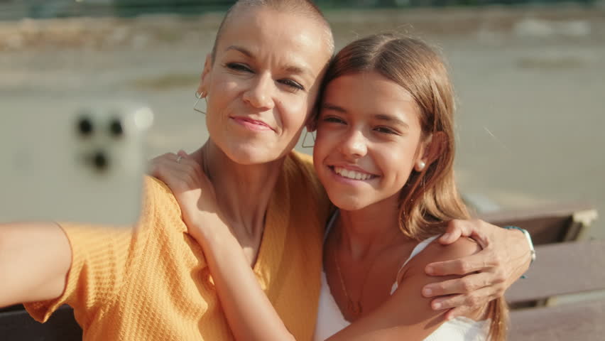 A mother with short hair and her daughter sit on a bench, happily browsing a phone together in a park.
