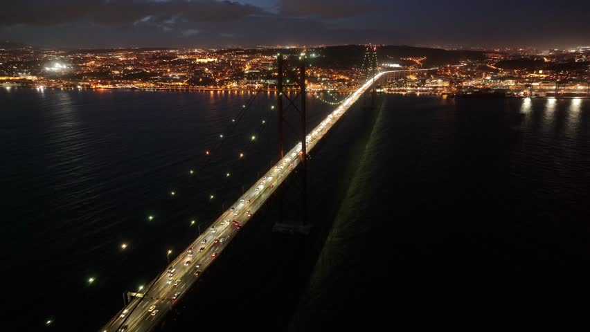 Night Scape Of 25 Of April Bridge At Lisbon In Lisbon District Portugal. Illuminated Downtown. Freeway Road Landscape. 25 Of April Bridge At Lisbon In Portugal. Cable Stayed Bridge Scenery.