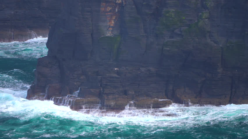 Powerful ocean waves crash against the base of the Cliffs of Moher, sending white spray into the air. The rugged coastline and towering cliffs showcase the untamed beauty of Ireland’s west coast.