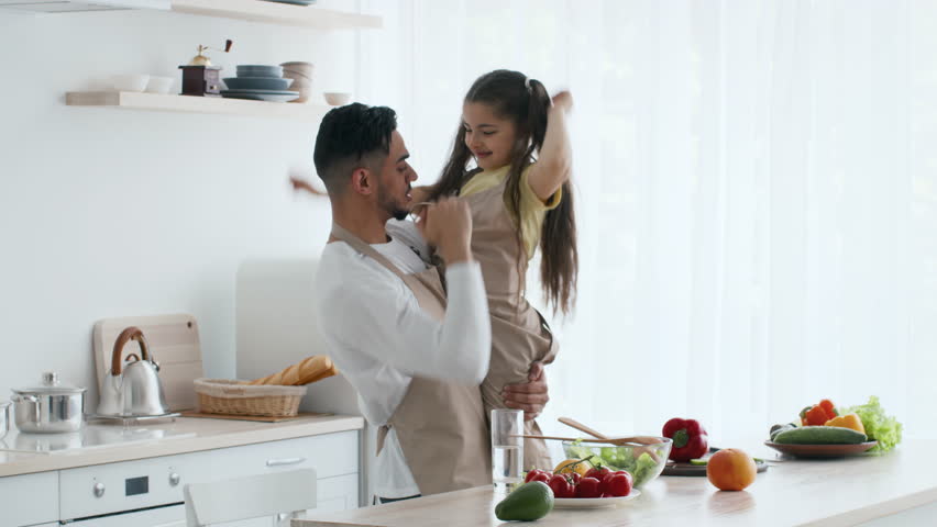 A joyful father lifts his daughter in the kitchen as they prepare a meal. The space is filled with fresh vegetables and fruits, creating a warm and inviting atmosphere. - Powered by Shutterstock - Get 15% off with code: PIKWIZARD15