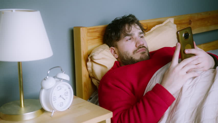Young man in red sweater lying in bed, holding smartphone with guilty expression, ignoring alarm clock, late morning procrastination, digital distraction