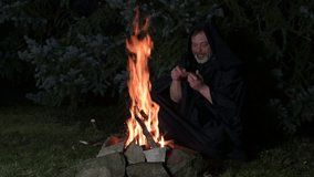 An elderly monk in a black robe counts money from his wallet while sitting by a fire at night in the woods. - Powered by Shutterstock - Get 15% off with code: PIKWIZARD15