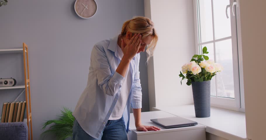 Tired overworked woman feeling dizzy and suffering from terrible headache, going to loose consciousness, posing in living room near window, being exhausted after working at laptop for long time