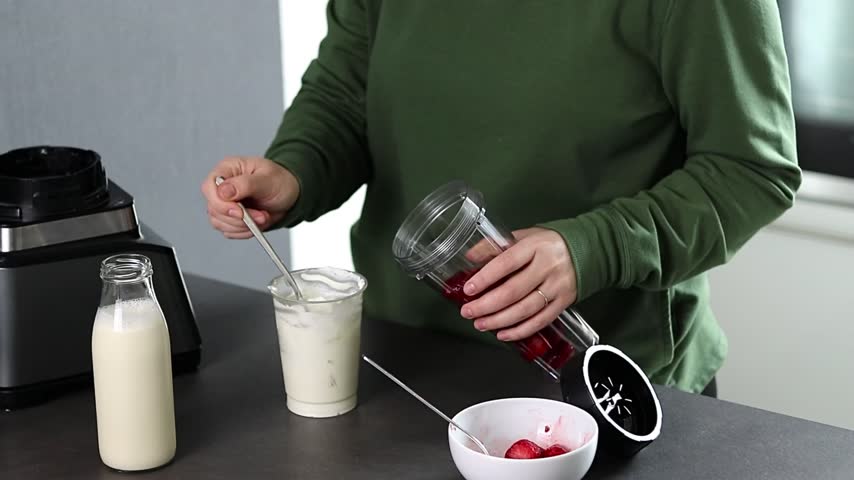 Close up of a woman in green sweater preparing fresh strawberries smoothie with yogurt and milk in the kitchen 