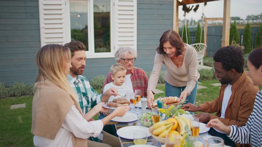 Camera view of family sitting together outside at table. Grandmother bringing plate with food to table. People starting eating. Visiting relatives in village or countryside. House in background.