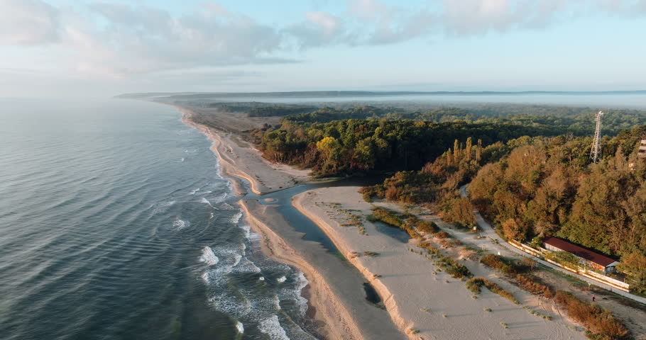 Aerial shot Kamchia River estuary meeting Black Sea Bulgaria. Greenery surrounds winding waterway forming scenic contrast golden sand deep blue sea. Coastal landscape highlights untouched beauty.