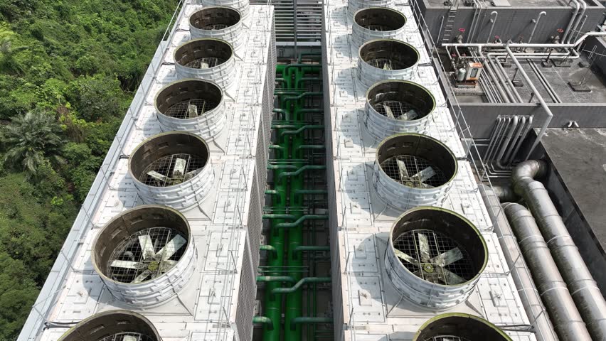 Aerial view of large industrial cooling towers with ventilation fans and green insulated piping system on building rooftop.