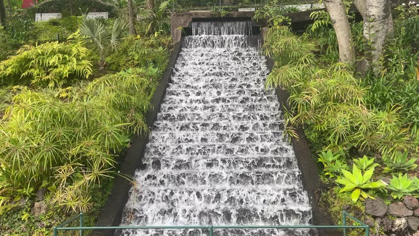 A stunning artificial waterfall cascades down moss-covered rocks at Monte Palace Botanical Garden, creating a tranquil oasis surrounded by exotic plants. 