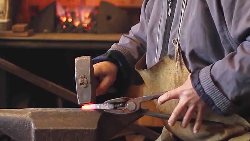 Blacksmith at work, close-up of man hitting a red-hot iron with a hammer on the anvil. Blacksmith forging red-hot metal bar in smithy