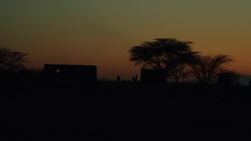 Kenya, Loima - September 9, 2023: Boy plays and jumps around man sitting in chair. View of houses in settlement in savannah and beautiful landscape with mountains silhouetted against evening sky