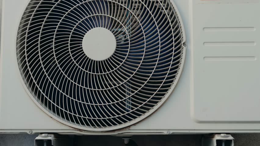 Close-up of the exterior unit of an air conditioner. The fan is visible through the circular grill. The unit is white and mounted on a wall. The video evokes feelings of comfort and relief,