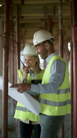 Professional architects wearing safety gear collaborating and reviewing blueprints at active construction site, communicating via walkie talkie with focused teamwork
