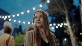 A young woman smiles gently, illuminated by string lights at a city night scene, exuding a serene vibe. Young Woman Enjoying a Night Out in the City Under Bright Lights - Powered by Shutterstock - Get 15% off with code: PIKWIZARD15