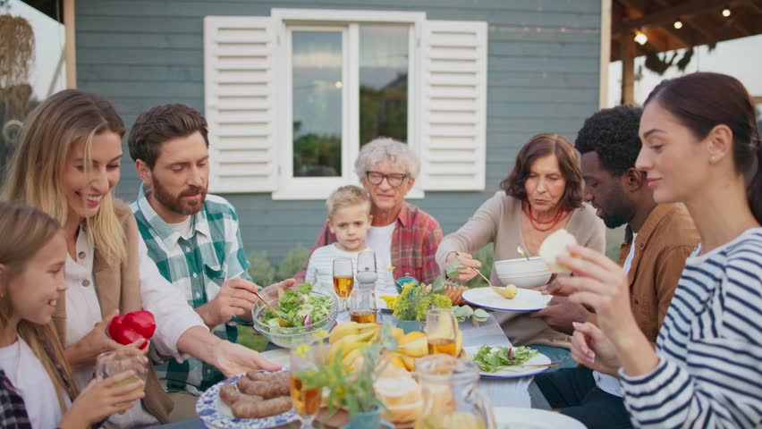 People having nice lunch together outside of house on green lawn. Enjoying warm weather. Placing food on dishes. Enjoying meal in cozy family gathering outside. Sunny warm weather.