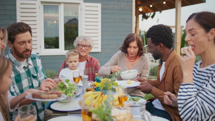 People having nice lunch together outside of house on green lawn. Enjoying warm weather. Placing food on dishes. Enjoying meal in cozy family gathering outside. Sunny warm weather.