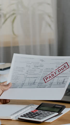 Vertical rear closeup of black male homeowner sitting at desk, holding overdue utilities invoice, using calculator to add up electricity and gas charges, while tracking household budget